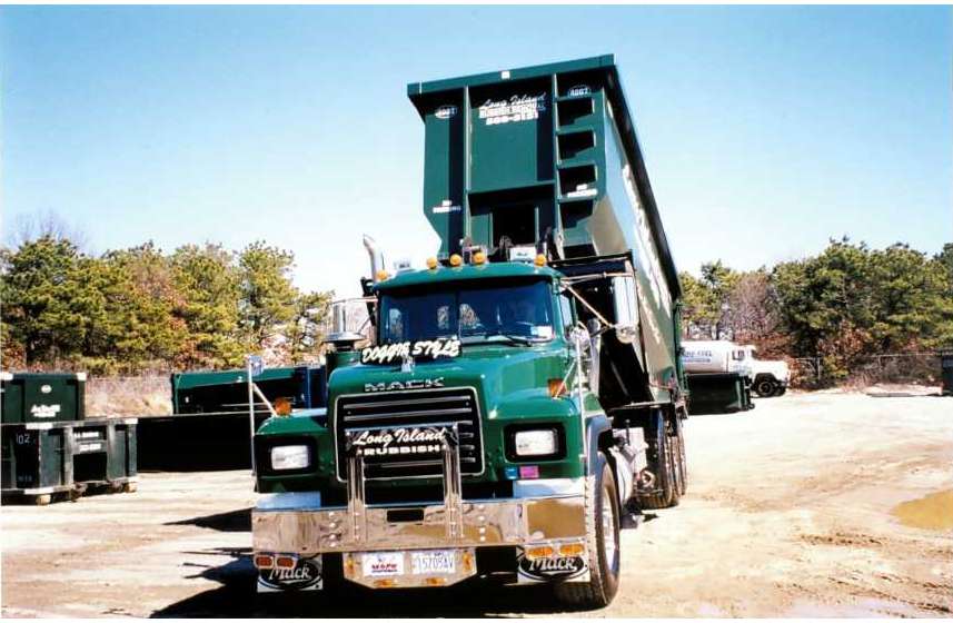 A green Mack dump truck with a raised bed, parked in a construction area with trees and containers in the background.